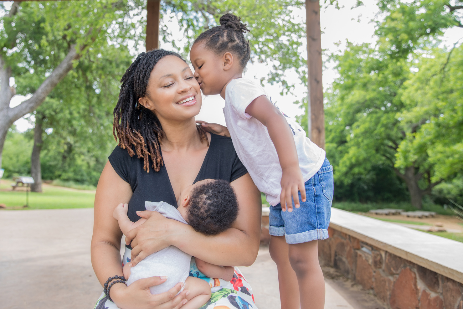 Toddler girl kisses mom while mom breastfeeds infant son